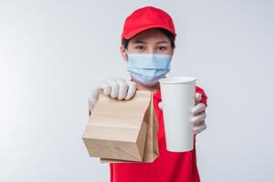 Image of a happy young delivery man in red cap blank t-shirt uniform face mask gloves standing with empty brown craft paper packet isolated on light gray background studio photo