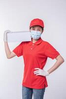 imagen de un joven repartidor consciente con gorra roja en blanco, camiseta uniforme, guantes de máscara facial de pie con una caja de cartón blanca vacía aislada en un estudio de fondo gris claro foto