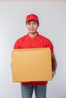 imagen de un joven y feliz repartidor con gorra roja, camiseta en blanco, uniforme de pie con una caja de cartón marrón vacía aislada en un estudio de fondo gris claro foto