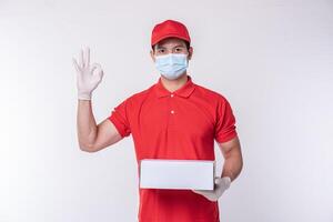 imagen de un joven repartidor consciente con gorra roja en blanco, camiseta uniforme, guantes de máscara facial de pie con una caja de cartón blanca vacía aislada en un estudio de fondo gris claro foto