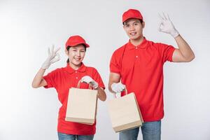 imagen de un joven repartidor feliz con gorra roja en blanco uniforme de camiseta de pie con un paquete de papel artesanal marrón vacío aislado en un estudio de fondo gris claro foto