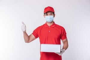 imagen de un joven repartidor consciente con gorra roja en blanco, camiseta uniforme, guantes de máscara facial de pie con una caja de cartón blanca vacía aislada en un estudio de fondo gris claro foto