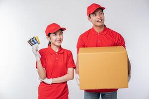 Image of a happy young delivery man with credit card in red cap blank t-shirt uniform standing with empty brown cardboard box isolated on light gray background studio photo