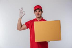 imagen de un joven y feliz repartidor con gorra roja, camiseta en blanco, uniforme de pie con una caja de cartón marrón vacía aislada en un estudio de fondo gris claro foto