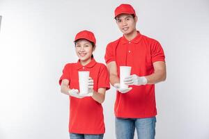 Image of a happy young delivery man in red cap blank t-shirt uniform standing with empty white paper cup isolated on light gray background studio photo