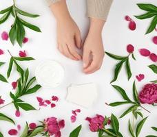 two female hands and a jar with thick cream and burgundy flowering peonies with green leaves photo
