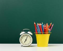 round alarm clock and multicolored pencils on the background of an empty green chalk board. Back to school photo