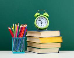 stack of books, a round alarm clock and multicolored pencils on the background of an empty green chalk board photo