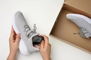 two female hands are holding a gray sports shoe over a white table, next to an open brown cardboard box photo