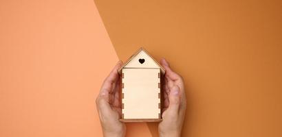 female hands folded to each other over a wooden miniature model house on a brown background. Real estate insurance concept photo