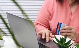 woman in a pink sweater makes purchases online with a laptop, a credit card in hand. Woman sitting at a white table and looking at the monitor photo