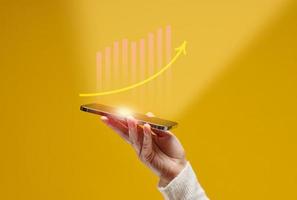 female hand holds a smartphone with holographic graphics on a yellow background. Performance growth, success photo