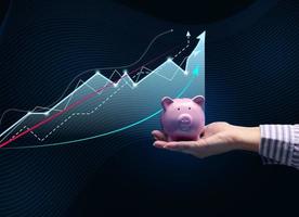 a female hand holds a pink ceramic piggy bank against the background of a growing holographic graph. Income growth concept, high margin in business, saving photo