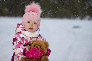 pequeño niña en invierno con un osito de peluche oso en el antecedentes de que cae nieve. niño en un invierno caminar. foto