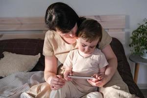 mamá y su pequeño hijo sentar en el cama y acecho dibujos animados en un teléfono inteligente el niño relojes un vídeo en un digital artilugio. auténtico estilo de vida, real familia hogar vida foto