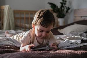 A two-year-old boy lies on a bed and watches cartoons on a smartphone photo