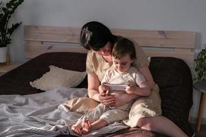 Mom and her little son sit on the bed and watching cartoons on a smartphone. The child watches a video on a digital gadget. Authentic lifestyle, real family home life photo