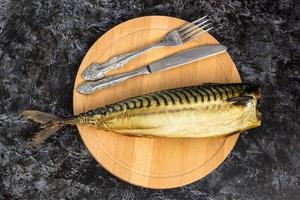 Smoked mackerel without head with fork knife cutting board on black background photo