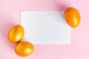 huevos de pascua dorados brillantes y tarjeta de felicitación en blanco sobre fondo rosa. foto