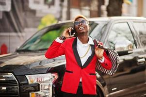 Fashion african american man model at red suit, with highlights hair posed against big suv black car and speaking on mobile phone. photo