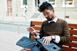Portrait of young stylish indian man model pose in street, sitting on bench with handbag. photo