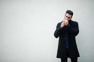 empresario del medio oriente usa abrigo negro y camisa azul, anteojos parados contra una pared blanca y fumando cigarrillos. foto