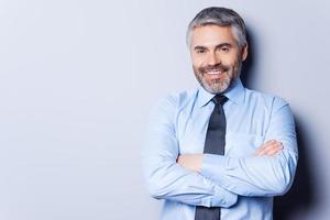 Confident and successful. Cheerful mature man in shirt and tie looking at camera and keeping arms crossed while standing against grey background photo