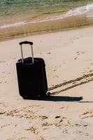 Black travel suitcase on sandy beach with turquoise sea background, summer holidays concept photo