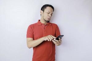 A dissatisfied young Asian man looks disgruntled wearing red t-shirt irritated face expressions holding his phone photo