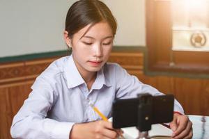 A women student with student suit is studying online from mobile phone at home with teacher, note teacher's explain knowledge on his book, education and online learning concept copy space for text photo