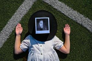 un niña en un blanco vestir es acostado en el fútbol americano campo. un mujer en un negro sombrero mentiras en el artificial césped en el Deportes campo. un cansado joven mujer es descansando en el Fresco aire. foto