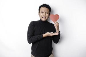 A portrait of an Asian man wearing a black shirt looks so confused while holding a red heart-shaped paper, isolated by a white background photo