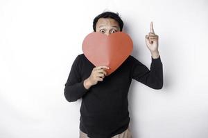 Portrait of a smiling Asian man holding a big red heart symbol pointing up at copy space isolated over white background photo