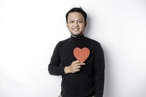 un retrato de un contento asiático hombre vistiendo un negro camisa, participación un rojo en forma de corazon papel aislado por blanco antecedentes foto