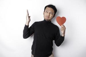 Confused Asian man holding a big red heart symbol pointing up at copy space isolated over white background photo