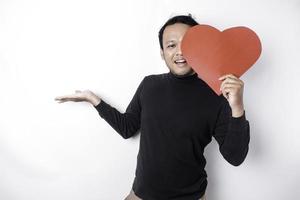 Excited Asian man wearing black shirt, pointing at the copy space beside him while holding a big red heart-shaped paper, isolated by white background photo