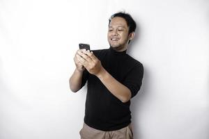 emocionado asiático hombre vistiendo negro camisa sonriente mientras participación su teléfono, aislado por blanco antecedentes foto