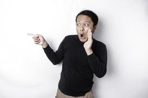 Shocked Asian man wearing black shirt pointing at the copy space beside him, isolated by white background photo