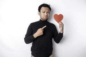 A portrait of an Asian man wearing a black shirt looks so confused while holding a red heart-shaped paper, isolated by a white background photo