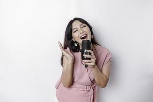 Portrait of carefree Asian woman, having fun karaoke, singing in microphone while standing over white background photo