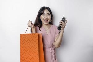 Portrait Asian happy beautiful young woman standing excited holding an online shopping bag and her smartphone, studio shot isolated on white background photo