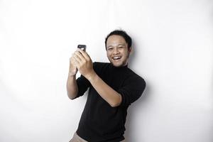 A young Asian man with a happy successful expression wearing black shirt and holding his phone, isolated by white background photo