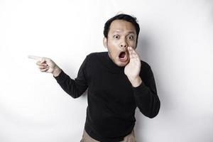 Shocked Asian man wearing black shirt pointing at the copy space beside him, isolated by white background photo