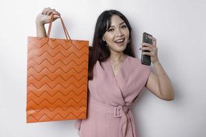 Portrait Asian happy beautiful young woman standing excited holding an online shopping bag and her smartphone, studio shot isolated on white background photo