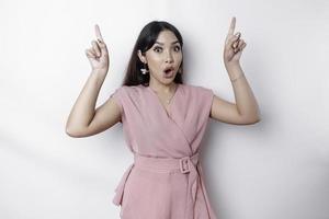 Shocked Asian woman dressed in pink, pointing at the copy space on top of her, isolated by white background photo