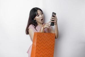 A young beautiful Asian woman standing surprised holding an online shopping bag and her smartphone, studio shot isolated on white background photo