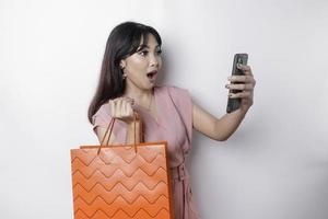 A young beautiful Asian woman standing surprised holding an online shopping bag and her smartphone, studio shot isolated on white background photo