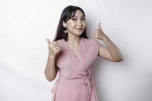 Excited Asian woman wearing pink blouse gives thumbs up hand gesture of approval, isolated by white background photo