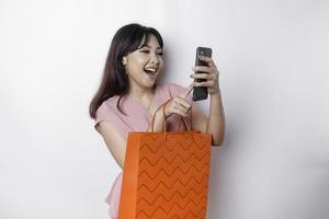 Portrait Asian happy beautiful young woman standing excited holding an online shopping bag and her smartphone, studio shot isolated on white background photo