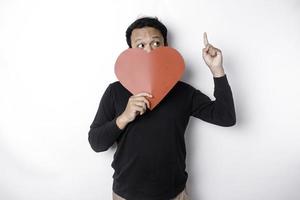 Portrait of a smiling Asian man holding a big red heart symbol pointing up at copy space isolated over white background photo